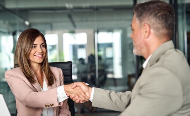 happy professional mature Latin businessman and businesswoman wearing suits shake hands sitting at table having partnership business contract agreement