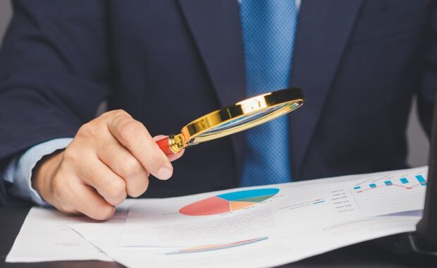 businessman holding a magnifying glass and looking at reading documents while sitting at a desk