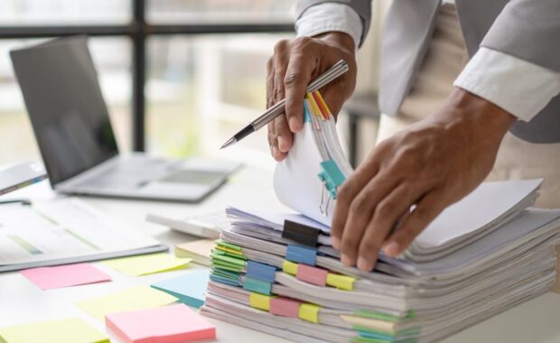 businessman working in stacks of papers searching for unfinished paperwork information on form check stack on table and checking financial papers in busy workload