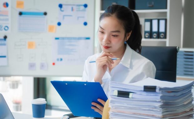 young Asian businesswoman works alone at a desk with a laptop, stack of papers, and documents