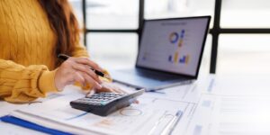 woman in a cozy yellow sweater is engaged in calculating figures on a calculator while reviewing financial reports next to a laptop