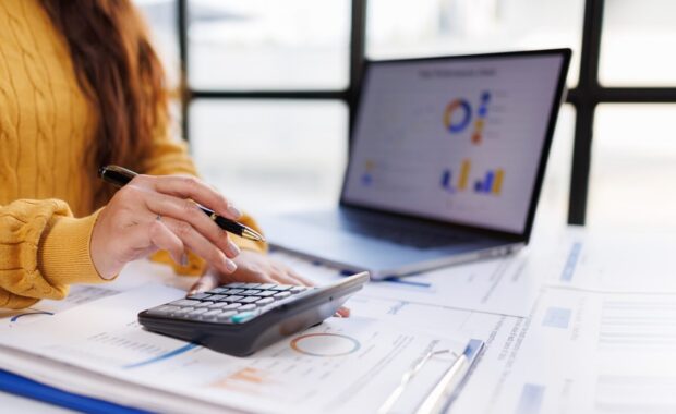 woman in a cozy yellow sweater is engaged in calculating figures on a calculator while reviewing financial reports next to a laptop