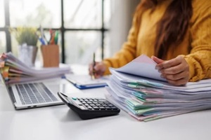 woman in a mustard sweater organizes colorful paperwork on her desk, while using a laptop, showcasing a busy and productive work environment